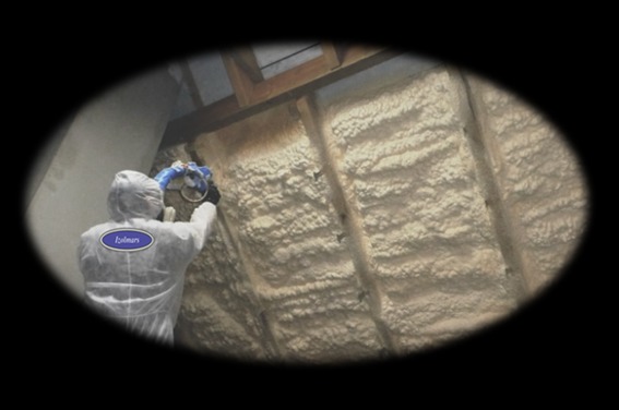 Pracownik w kombinezonie ochronnym aplikuje piankę izolacyjną na poddaszu, widoczne drewniane elementy konstrukcyjne dachu i logo firmy na kombinezonie.