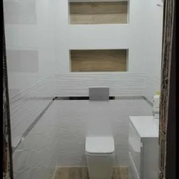 Nowoczesna łazienka z białymi płytkami, drewnopodobną podłogą i wnękami z drewnianym akcentem, widok na toaletę, umywalkę i fragment ściany z dekoracyjnymi liniami.