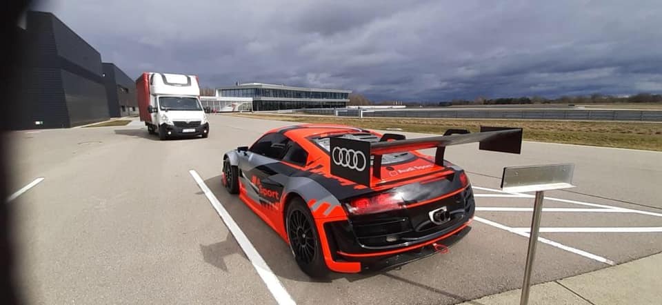 Czerwono-szary samochód wyścigowy Audi z dużym spojlerem na torze, w tle biała furgonetka i budynek z ciemną elewacją, pochmurne niebo.