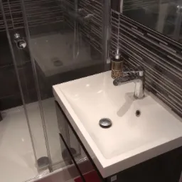 Nowoczesna łazienka z białą umywalką nablatową, chromowaną baterią i kabiną prysznicową z przezroczystego szkła, wykończona płytkami w odcieniach szarości i brązu, z widocznym gniazdkiem...