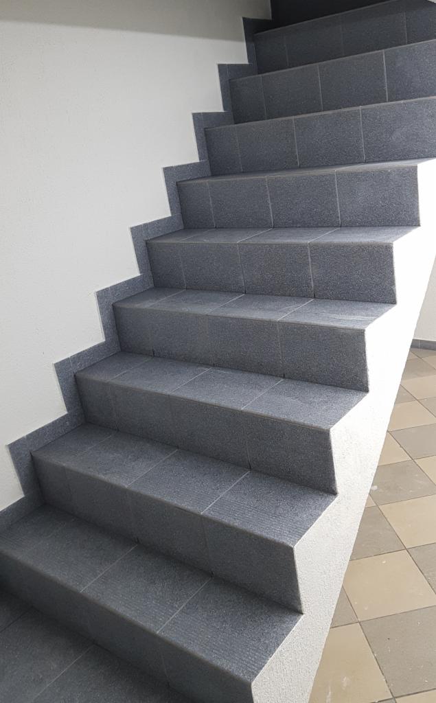 Schody wyłożone szarymi, kwadratowymi płytkami z delikatną fakturą, widoczne z dołu, z fragmentem podłogi w szachownicę w prawym dolnym rogu.