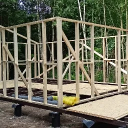 Budowa szkieletowej konstrukcji z drewna na fundamencie z izolacją, w tle las, pracownik montuje płytę OSB.