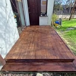 Wood is Life Tomasz Pykało - Ciemnobrązowy taras drewniany z dwoma stopniami, przylegający do białego budynku z ciemnymi drzwiami. Widok z góry, w słoneczny dzień.