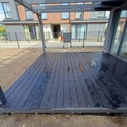 Wood is Life Tomasz Pykało - Nowoczesny taras z ciemnoszarej deski kompozytowej pod minimalistyczną pergolą. Widok na budynek mieszkalny w tle, zieleń i ogrodzenie. Świeżo po deszczu.