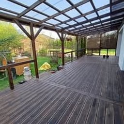 Wood is Life Tomasz Pykało - Zadaszony taras z brązową deską kompozytową, drewnianą konstrukcją i przezroczystym zadaszeniem. Widok na zielony ogród z elementami wyposażenia.