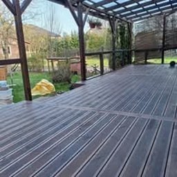 Wood is Life Tomasz Pykało - Zadaszony taras z desek kompozytowych w odcieniach szarości, z widoczną konstrukcją drewnianą i elementami ogrodu w tle, uchwycony w słoneczny dzień.