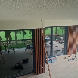 Wood is Life Tomasz Pykało - Montaż drewnianej zabudowy balkonu z lamelami. Widoczne narzędzia i częściowo zamontowane elementy. Prace w toku, z widokiem na zewnątrz przez duże okna.