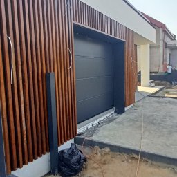 Wood is Life Tomasz Pykało - Nowoczesna brama garażowa w kolorze antracytu z drewnianą okładziną lamelową. Widoczne elementy wykończeniowe i fragment posesji w trakcie prac.