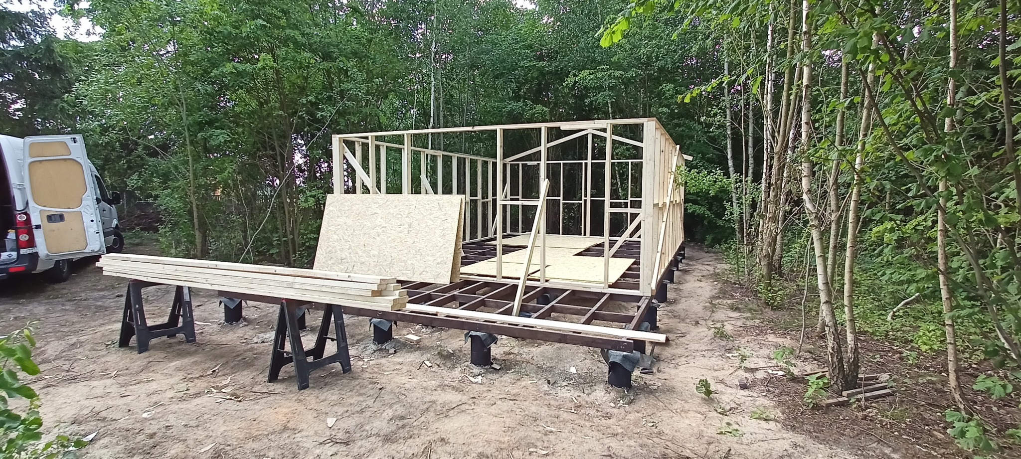 Konstrukcja szkieletowa budynku w trakcie budowy, widoczne elementy drewniane, płyty OSB, podpora na regulowanych słupkach, tło leśne, biały bus w tle.