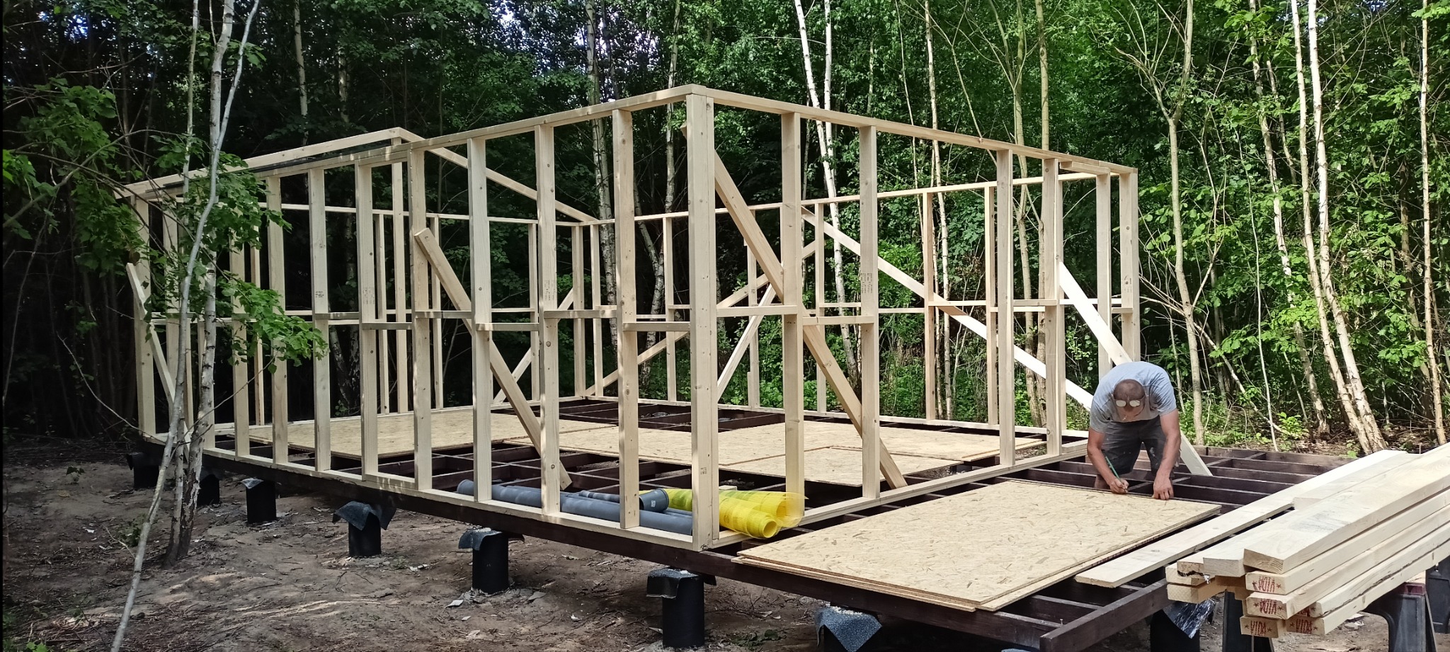 Budowa szkieletowej konstrukcji z drewna na fundamencie z izolacją, w tle las, pracownik montuje płytę OSB.