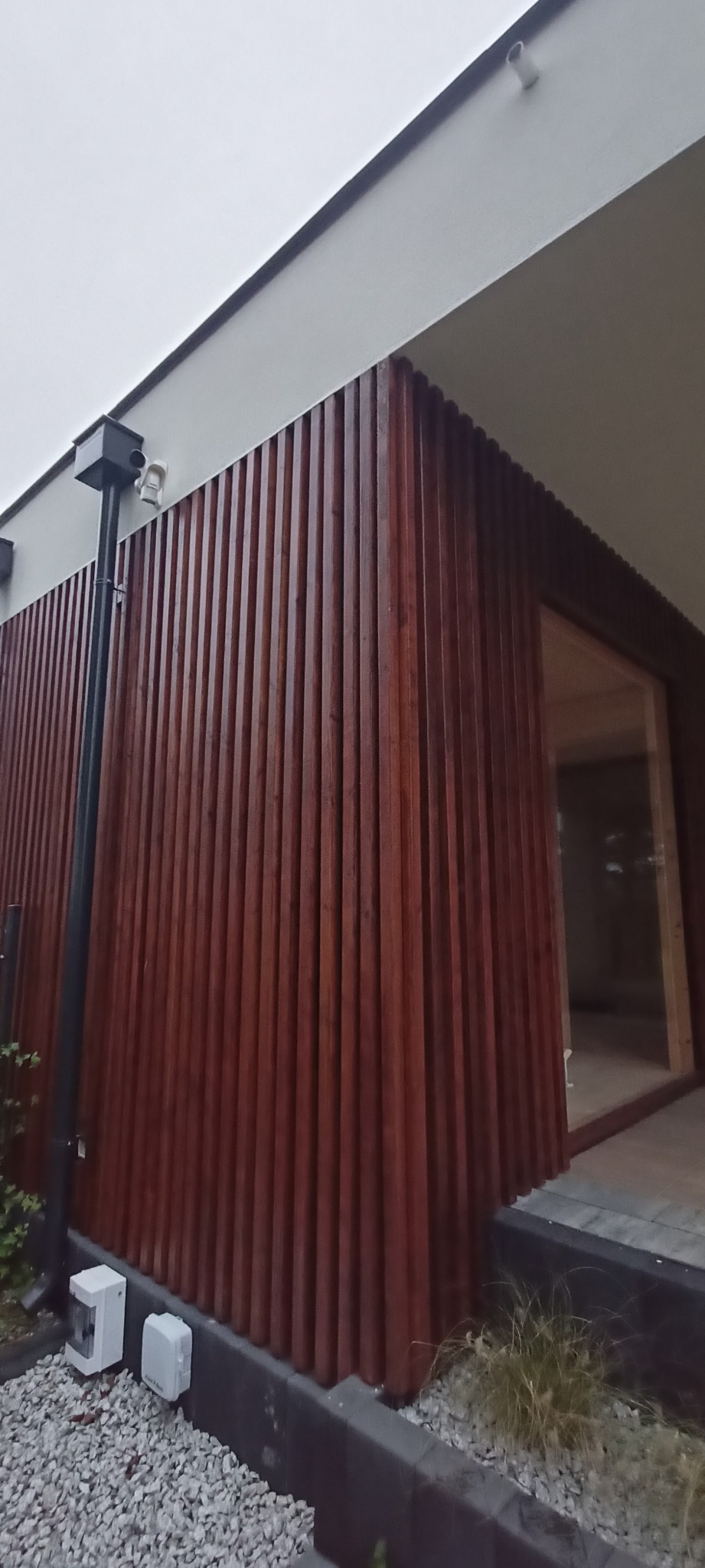 Nowoczesna elewacja z drewnianych paneli w Siemiatyczach. Pionowe deski w ciepłym odcieniu brązu tworzą unikalny wzór. Widoczny fragment budynku z oknem i elementami instalacji.