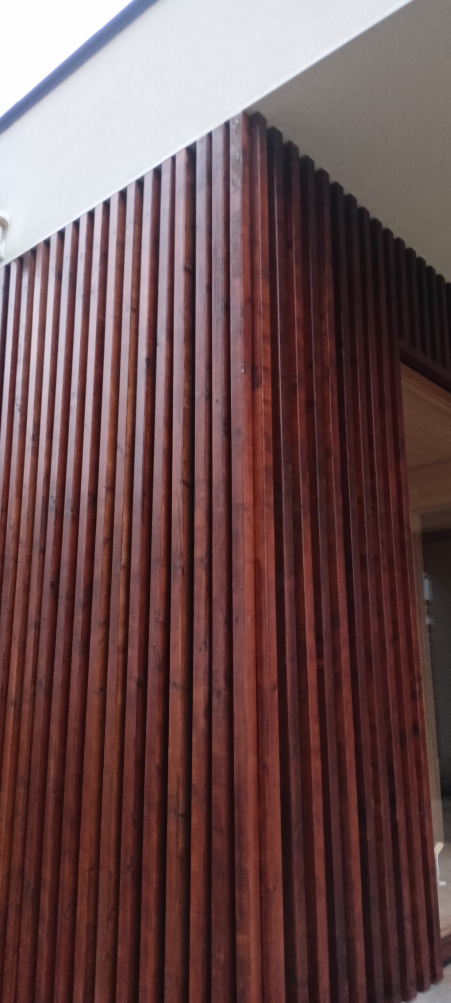 Drewniana elewacja budynku z pionowymi listwami w ciepłym odcieniu brązu, tworząca nowoczesny i elegancki wygląd. Widoczny fragment dachu i drzwi.