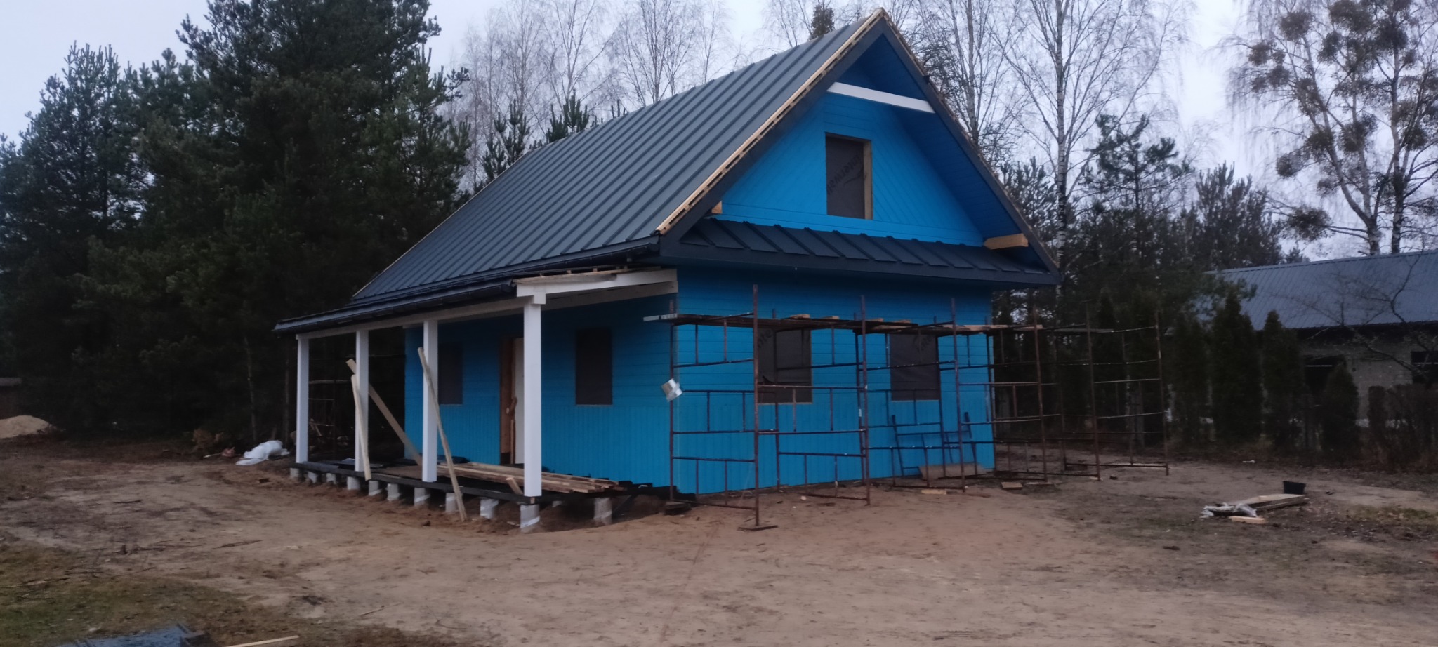 Niebieski domek z szarą blachodachówką w trakcie budowy, z widocznym rusztowaniem i elementami konstrukcyjnymi werandy. Otoczenie leśne.