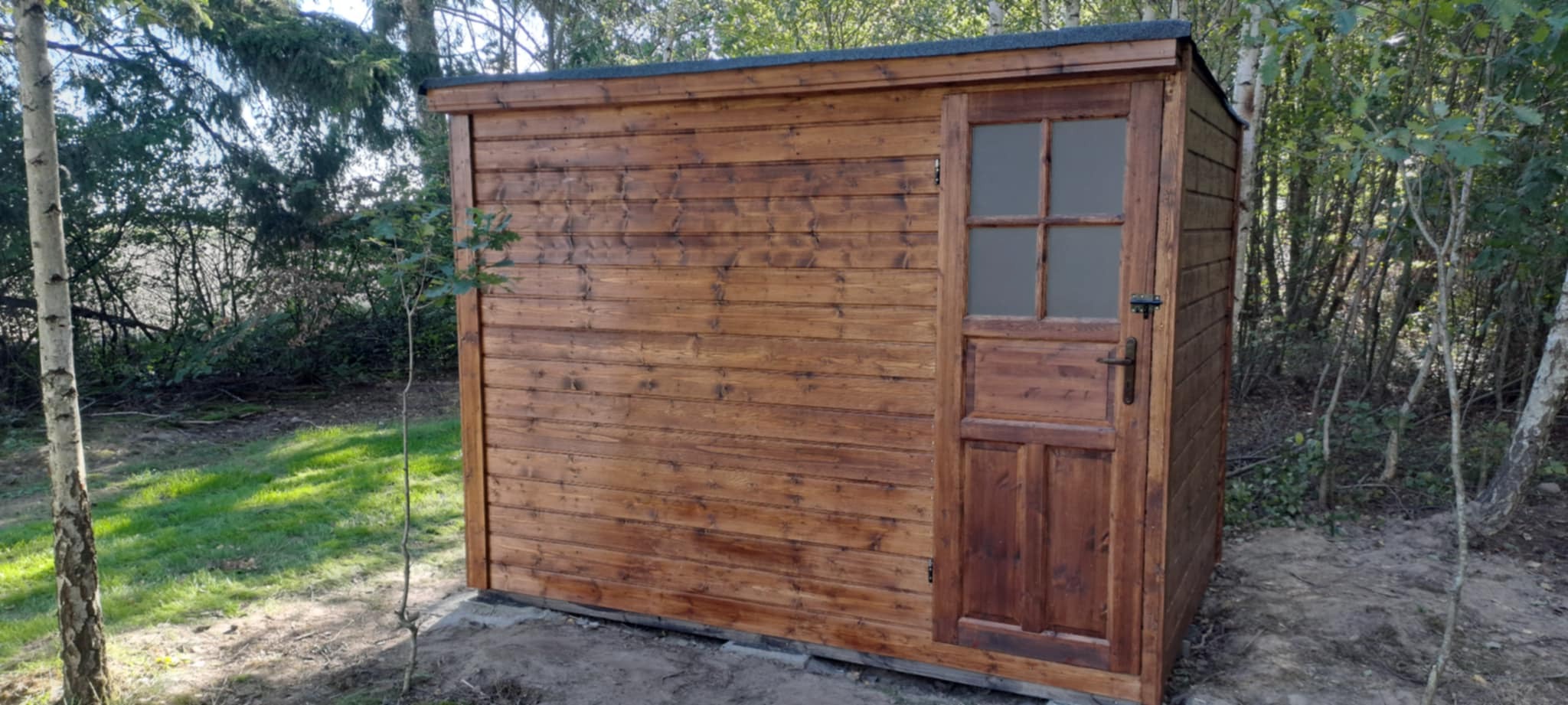 Drewniana altana z pojedynczymi drzwiami z szybą, pomalowana na brązowo, otoczona zielenią drzew i krzewów. Widok z boku, w tle las.