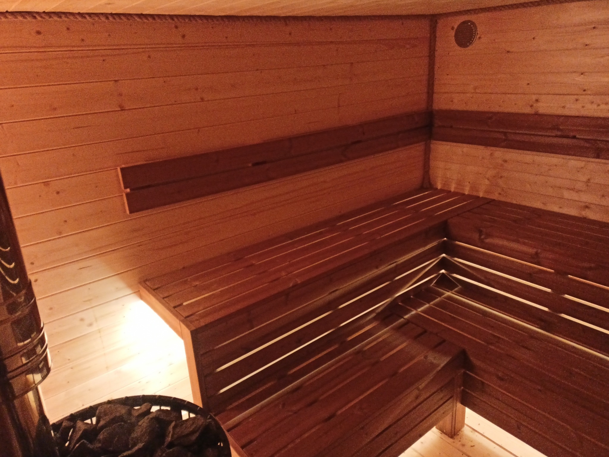 Wnętrze sauny wykończone drewnem, z piecem na kamienie w rogu. Widoczne drewniane ławki i oświetlenie. Ciepła kolorystyka drewna tworzy relaksującą atmosferę.
