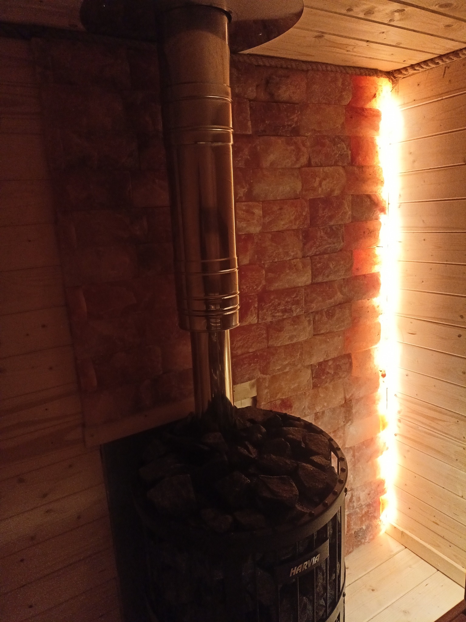 Wnętrze sauny z piecem Harvia wypełnionym kamieniami, kominem i ścianą z cegieł solnych podświetloną od tyłu, tworząc ciepłą atmosferę.