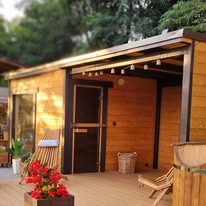 Drewniana sauna ogrodowa z przeszklonymi drzwiami, oświetlona girlandą żarówek, z elementami dekoracyjnymi na tarasie. Ujęcie z boku, w ciepłym oświetleniu.