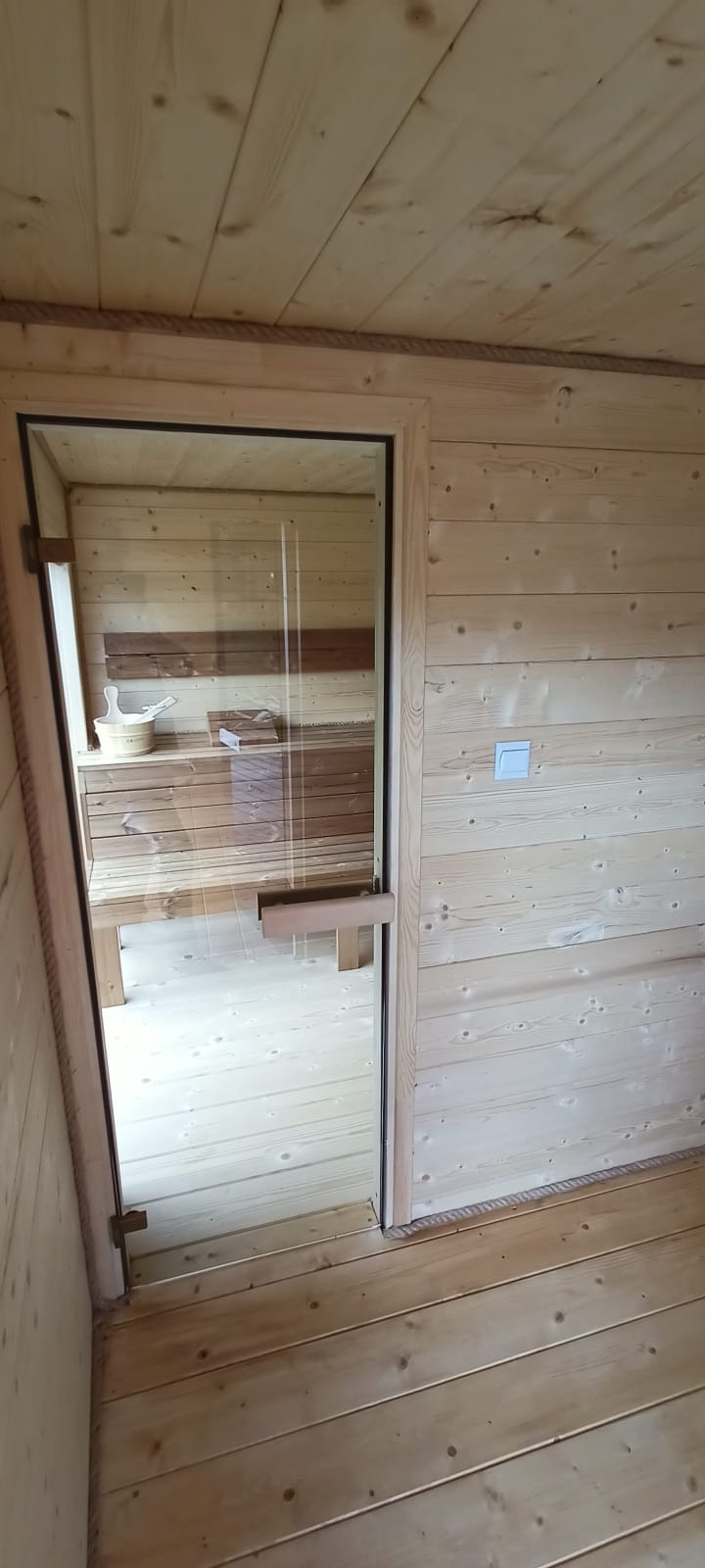 Wnętrze sauny z drewnianym wykończeniem, widok na ławki przez szklane drzwi z drewnianą klamką. Na ścianie widoczny włącznik światła. Całość w jasnej tonacji.