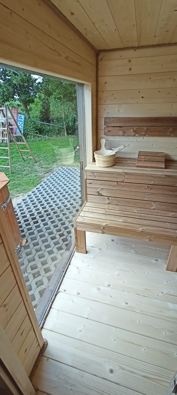 Jasne, drewniane wnętrze sauny z ławkami, wiadrem i chochlą. Przez okno widoczny trawnik z ażurowymi płytami i plac zabaw.