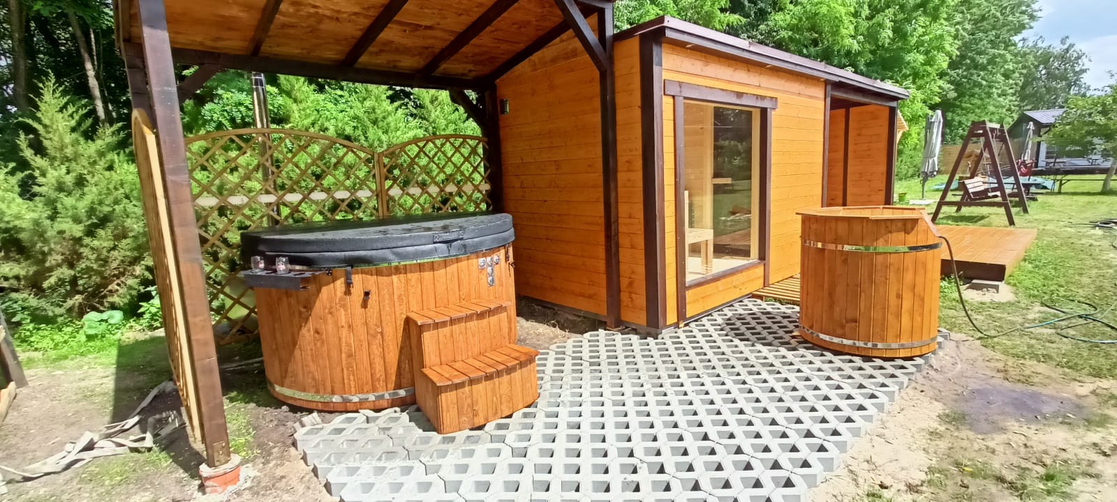 Drewniana sauna z oknem, beczka z wodą i jacuzzi pod zadaszeniem na betonowej nawierzchni. Całość w ogrodzie, w słoneczny dzień.