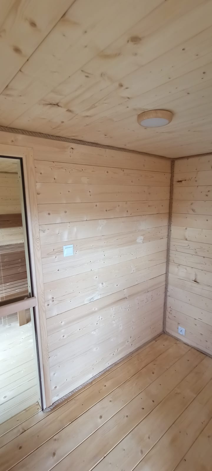 Wnętrze sauny wykończone jasnym drewnem, z widocznymi deskami na ścianach, podłodze i suficie. Dekoracyjne liny wzdłuż krawędzi. Widoczne gniazdka elektryczne i oświetlenie.