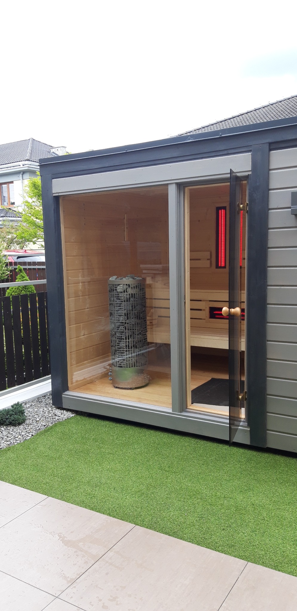 Nowoczesna sauna zewnętrzna z przeszklonymi ścianami, widoczny piec z kamieniami i drewniane ławy, otoczona sztuczną trawą i płytami tarasowymi.