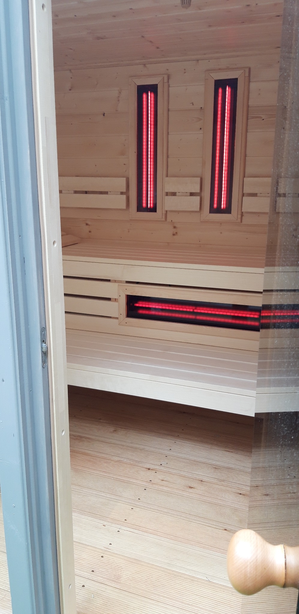 Wnętrze sauny z jasnego drewna z czerwonymi panelami grzewczymi. Widoczne ławki, podłoga z desek i fragment otwartych drzwi.