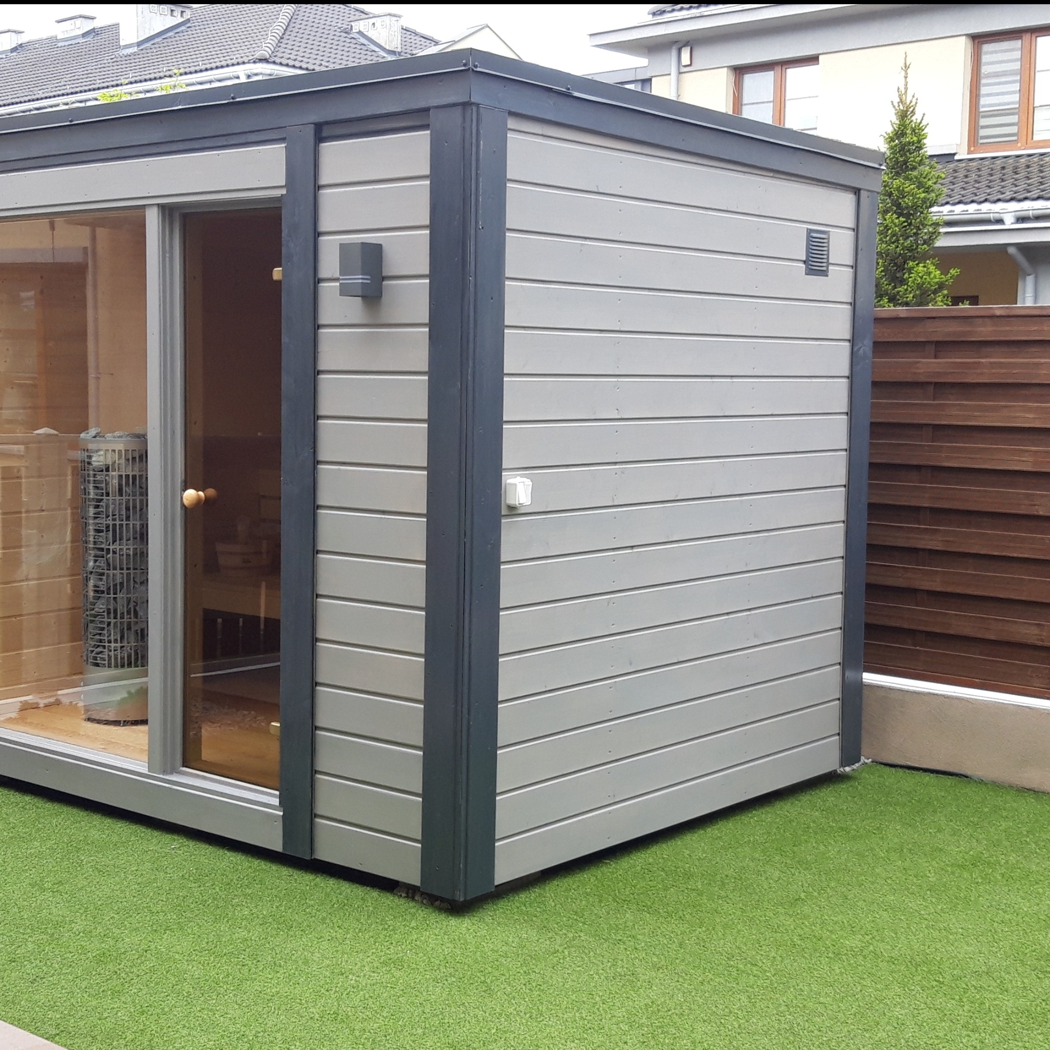 Nowoczesna sauna zewnętrzna z przesuwnymi drzwiami w Siemiatyczach. Szara drewniana konstrukcja na sztucznej trawie, widoczne wnętrze z piecem.