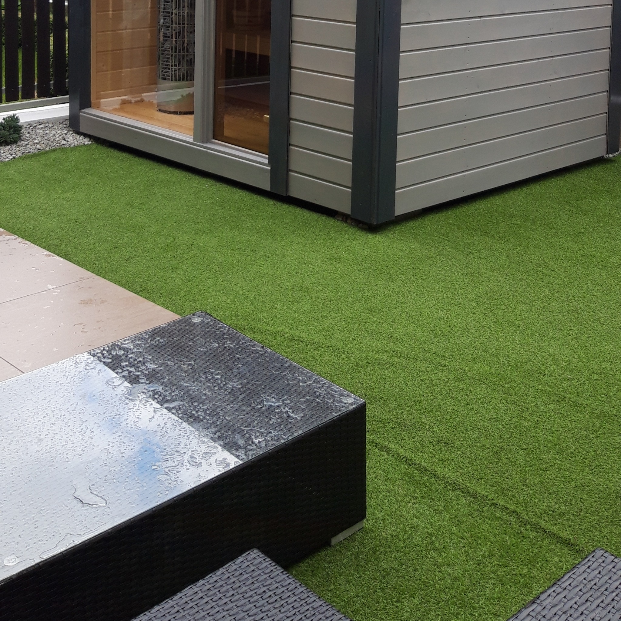 Nowoczesna sauna ogrodowa z szarymi panelami, widoczna przez okno konstrukcja z kamieni. Obok sztuczna trawa i meble ogrodowe z kroplami deszczu.