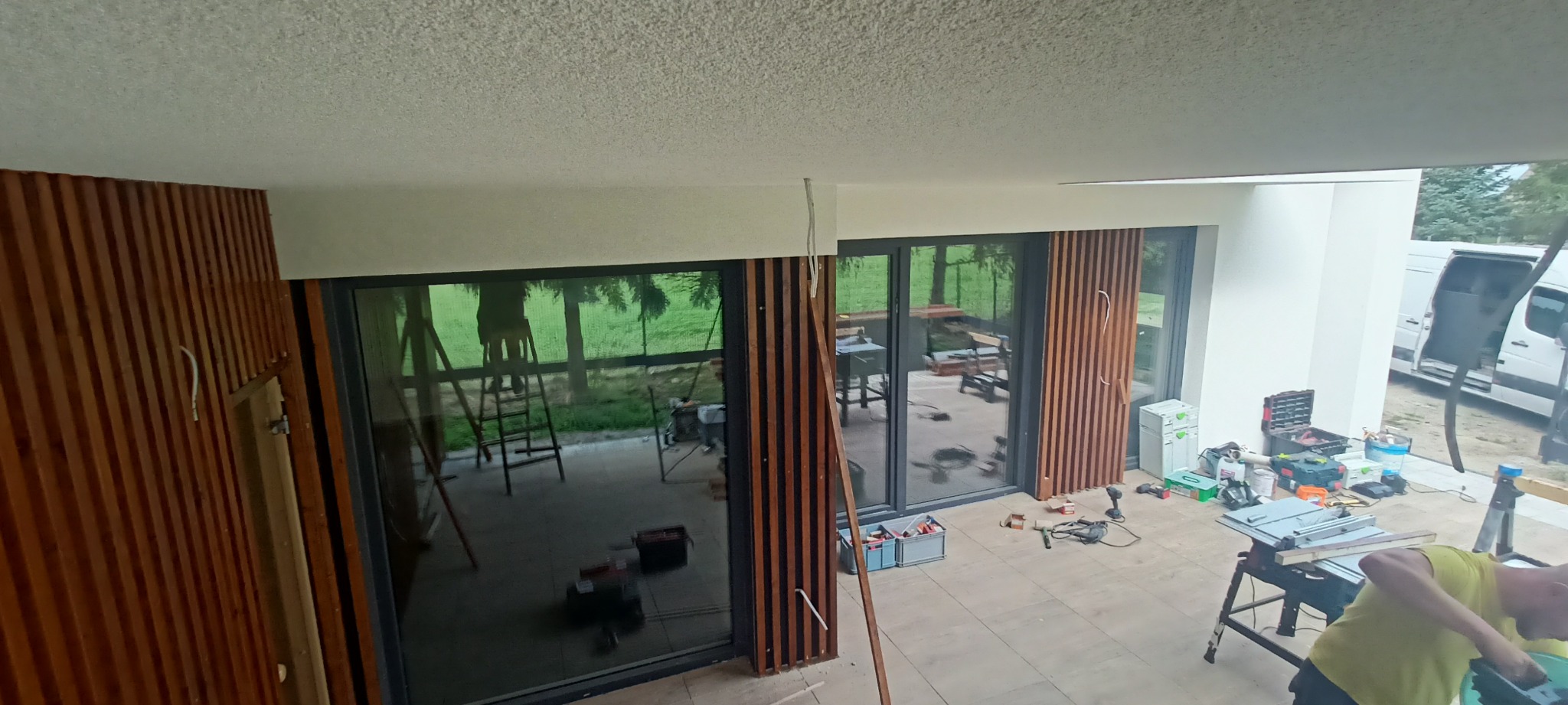 Montaż drewnianej zabudowy balkonu z lamelami. Widoczne narzędzia i częściowo zamontowane elementy. Prace w toku, z widokiem na zewnątrz przez duże okna.