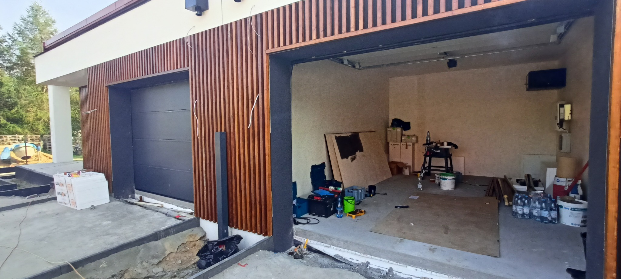 Nowoczesna elewacja z drewnianymi lamelami przy garażu w trakcie budowy. Widoczne materiały budowlane i narzędzia wewnątrz. Ujęcie z zewnątrz, częściowo ukończone.