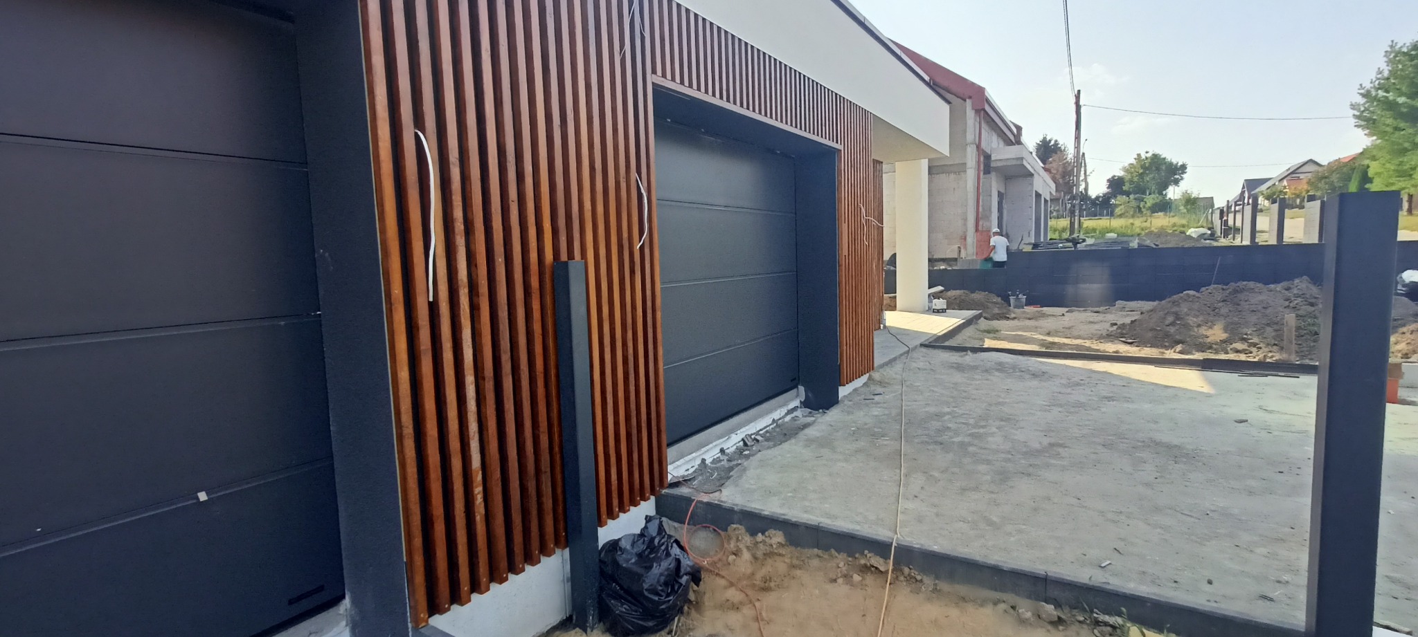 Nowoczesna brama garażowa w kolorze antracytu z drewnianą okładziną lamelową. Widoczne elementy wykończeniowe i fragment posesji w trakcie prac.