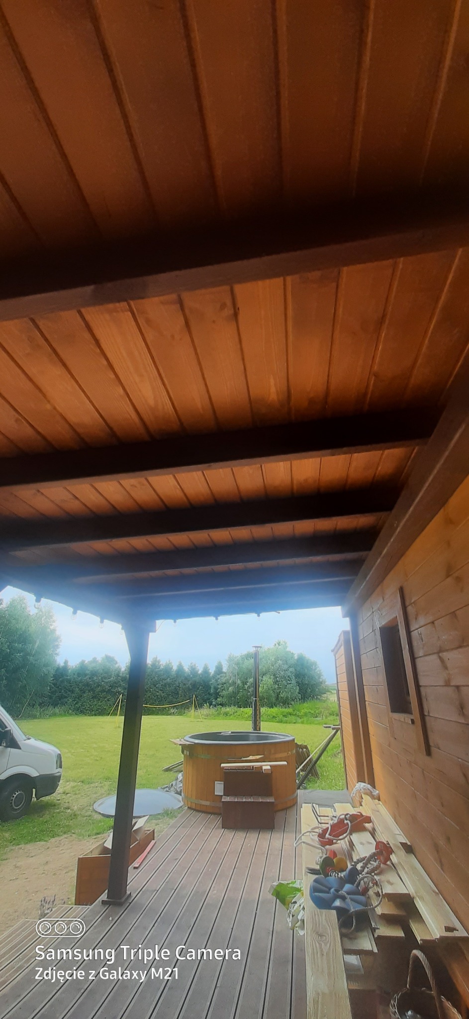 Drewniana konstrukcja z zadaszeniem i balia ogrodowa w tle. Widok z dołu na drewniany sufit i balie, otoczone zielenią. Częściowo widoczny samochód dostawczy.