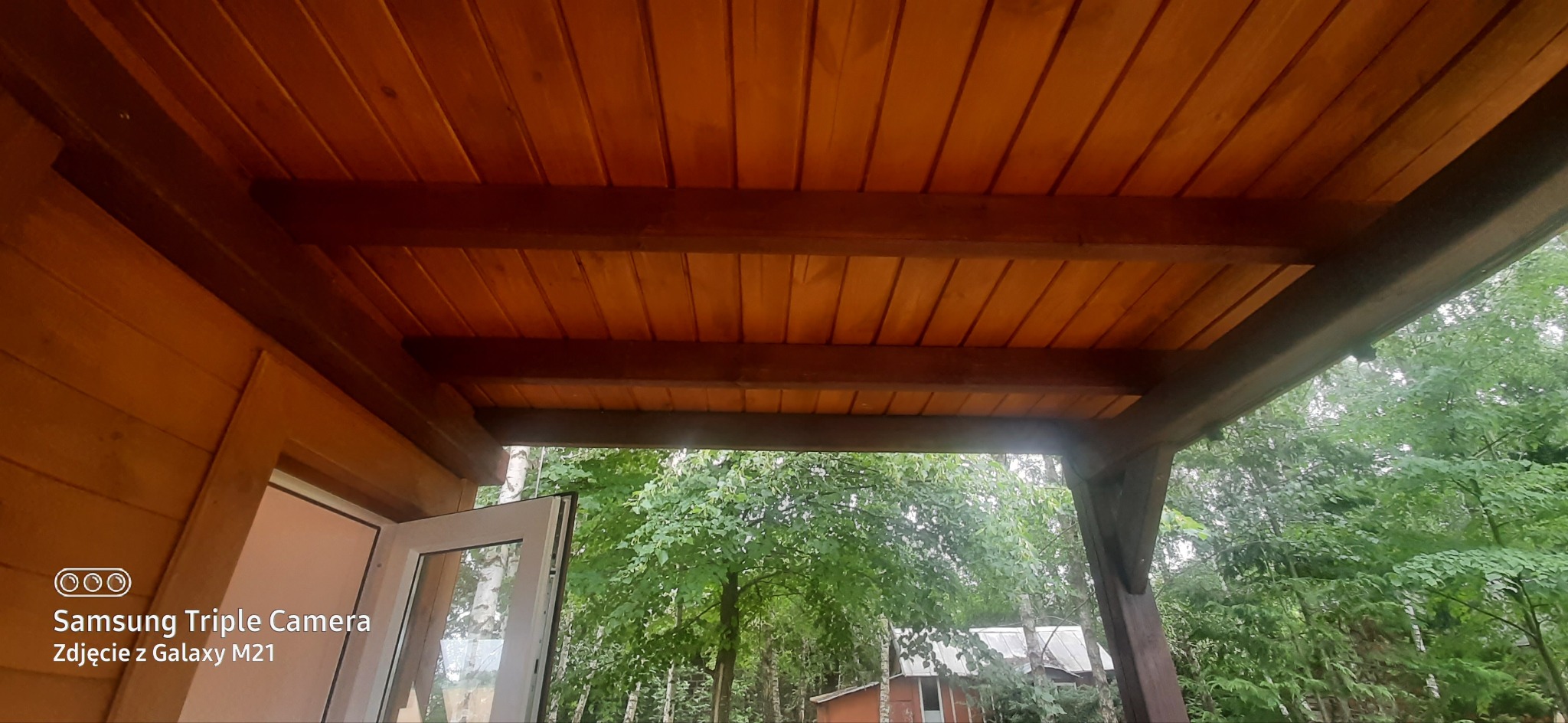Drewniana konstrukcja zadaszenia, widok od dołu, z widokiem na zieleń drzew. Elementy drewniane w ciepłych odcieniach brązu. Widoczny fragment budynku z oknem.