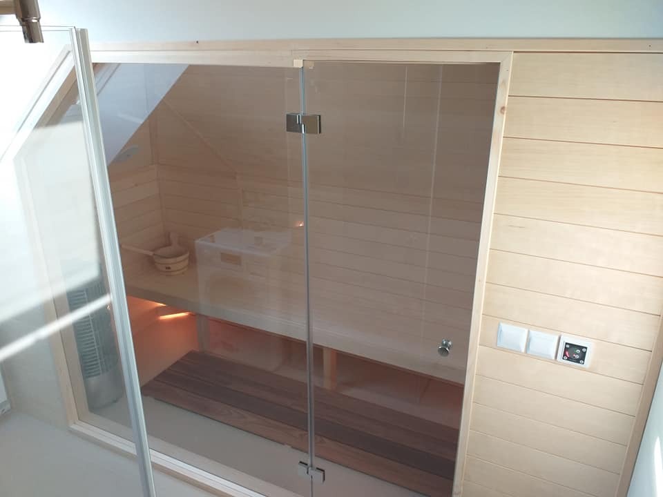 Jasna, drewniana sauna z przeszklonymi drzwiami, widoczne wnętrze z ławkami, oświetleniem i akcesoriami. Ujęcie z bliska, podkreślające detale wykończenia.