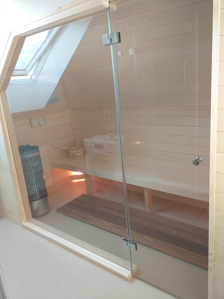 Jasna sauna w skandynawskim stylu z przeszklonymi drzwiami i drewnianym wykończeniem. Widoczny piec, ławki i okno dachowe.