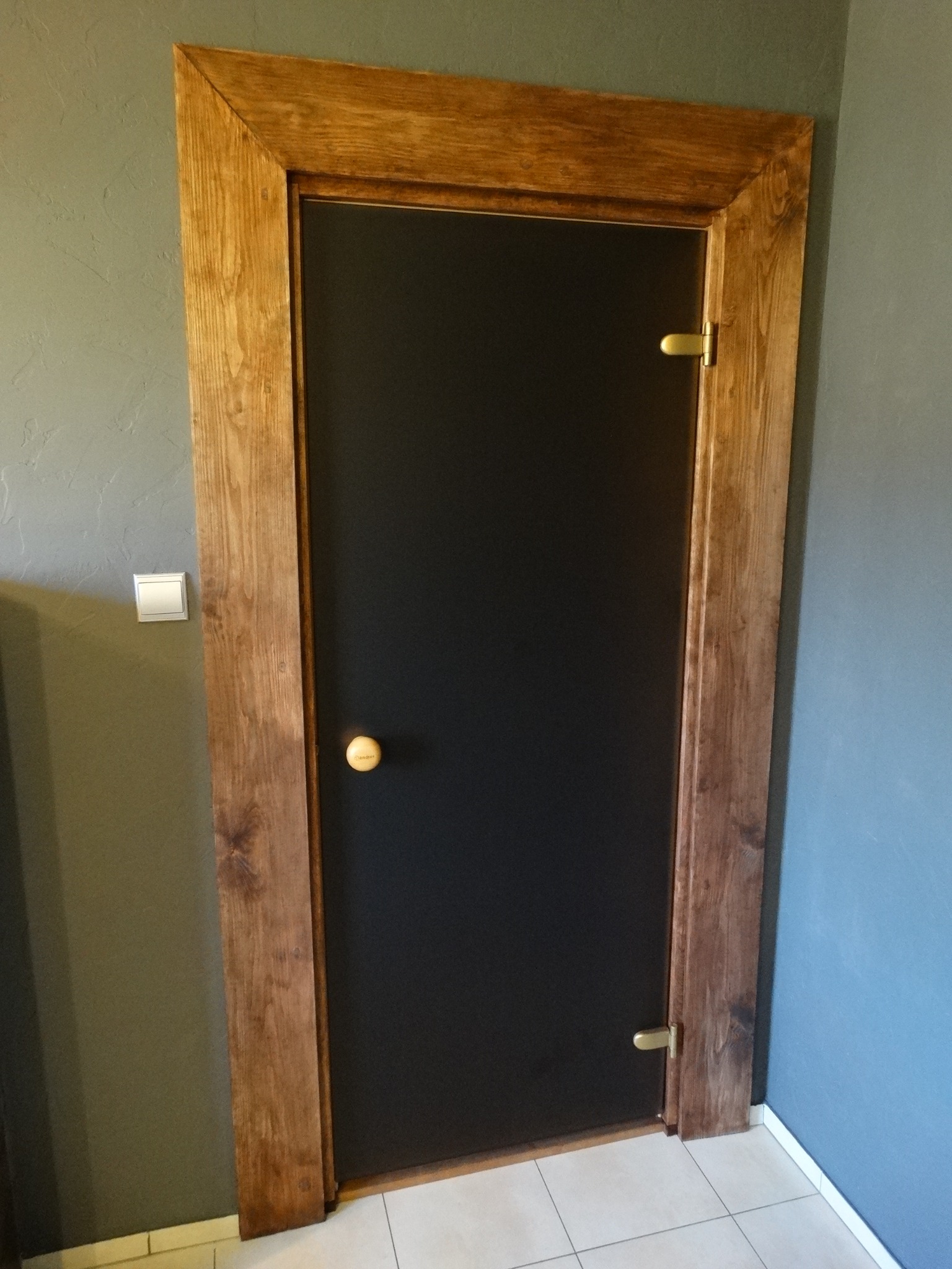 Ciemne drzwi z drewnianą ramą w saunie, widoczne detale zawiasów i klamki, płytki podłogowe w jasnym odcieniu, ściany w odcieniach szarości.