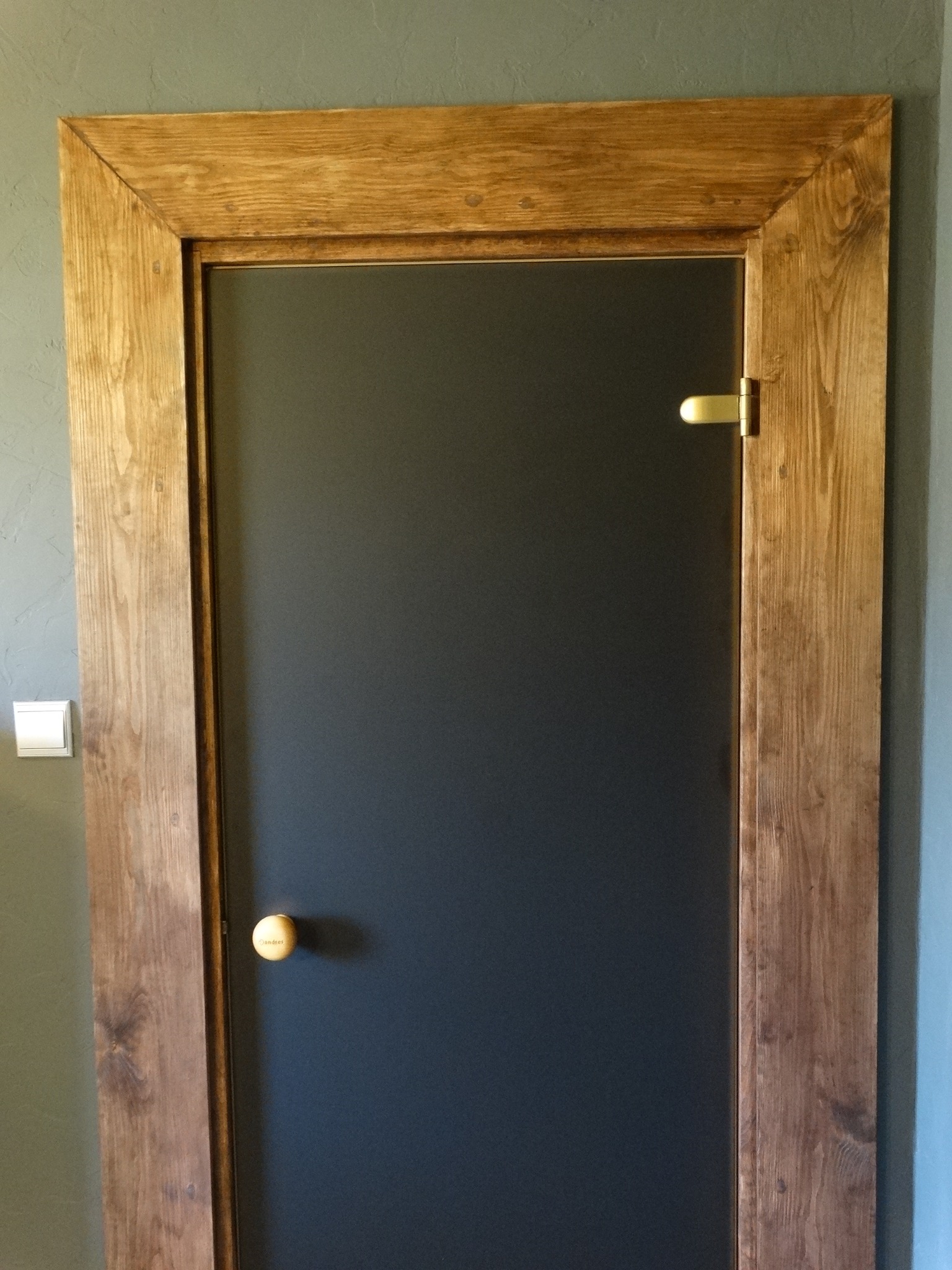 Ciemne drzwi z drewnianą ramą, widoczne zawiasy i okrągła klamka. Drewno ramy ma wyraźny rysunek słojów. Na ścianie obok drzwi widoczny włącznik światła.