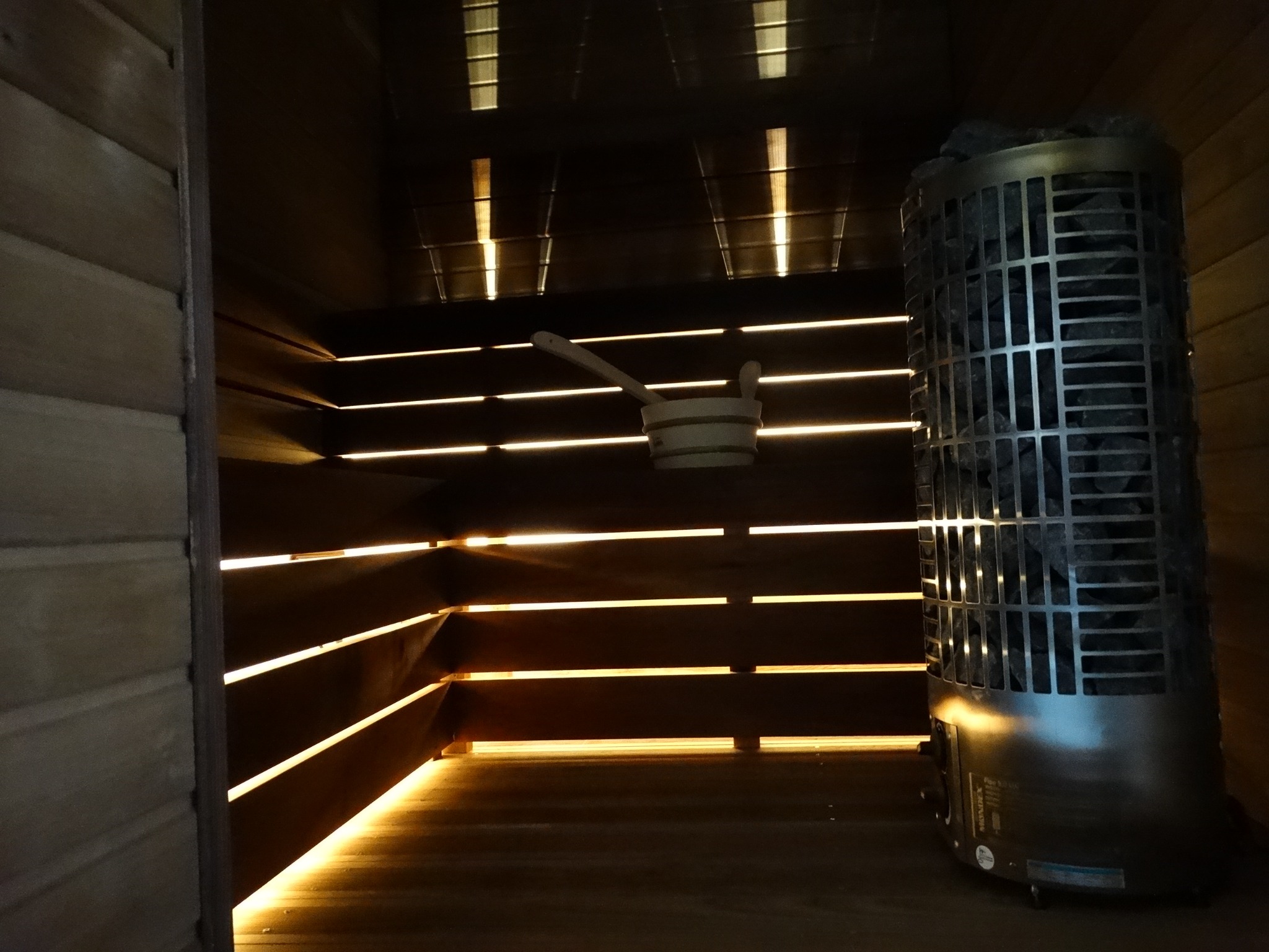 Wnętrze sauny z drewnianymi ławkami podświetlonymi ledami, piecem na kamienie i drewnianym czerpakiem z chochlą. Ciepłe światło tworzy relaksującą atmosferę.