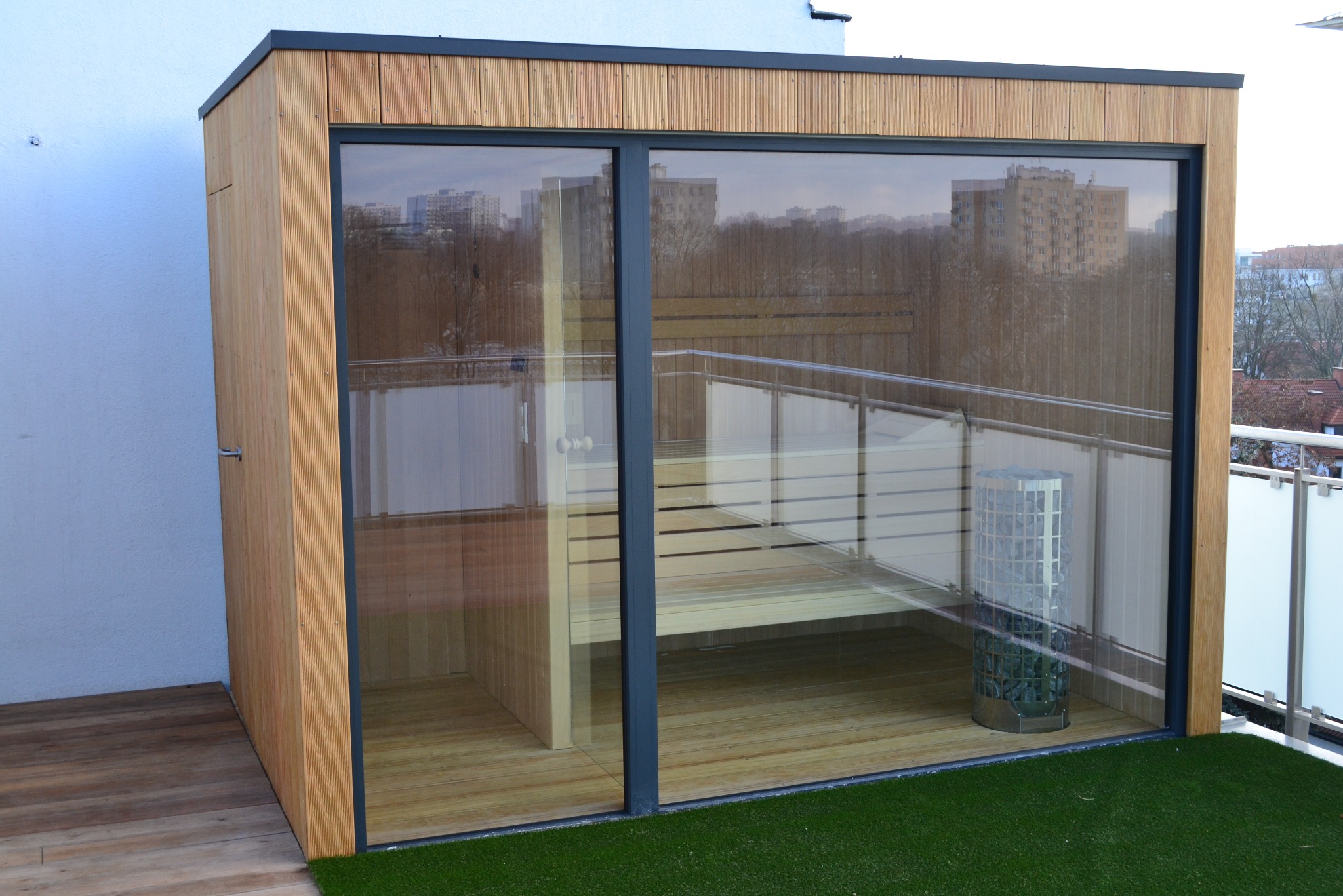 Nowoczesna sauna zewnętrzna z drewna, z dużym przeszkleniem, widoczna z zewnątrz na tle miejskiego krajobrazu. Na podłodze sztuczna trawa.