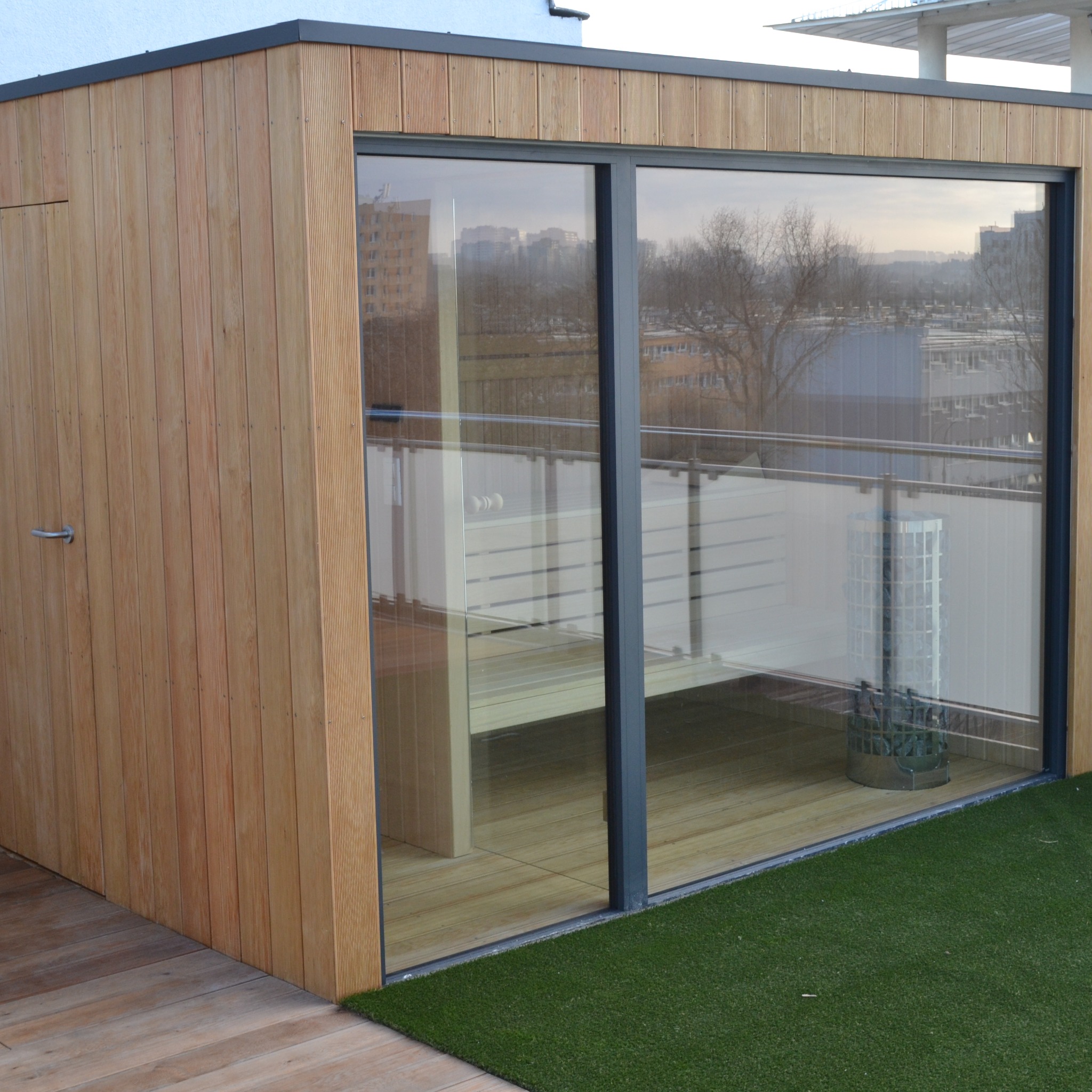 Nowoczesna sauna zewnętrzna z drewnianą elewacją i dużym oknem, widoczna z zewnątrz na tle miejskiego krajobrazu. Podłoga z desek, sztuczna trawa.