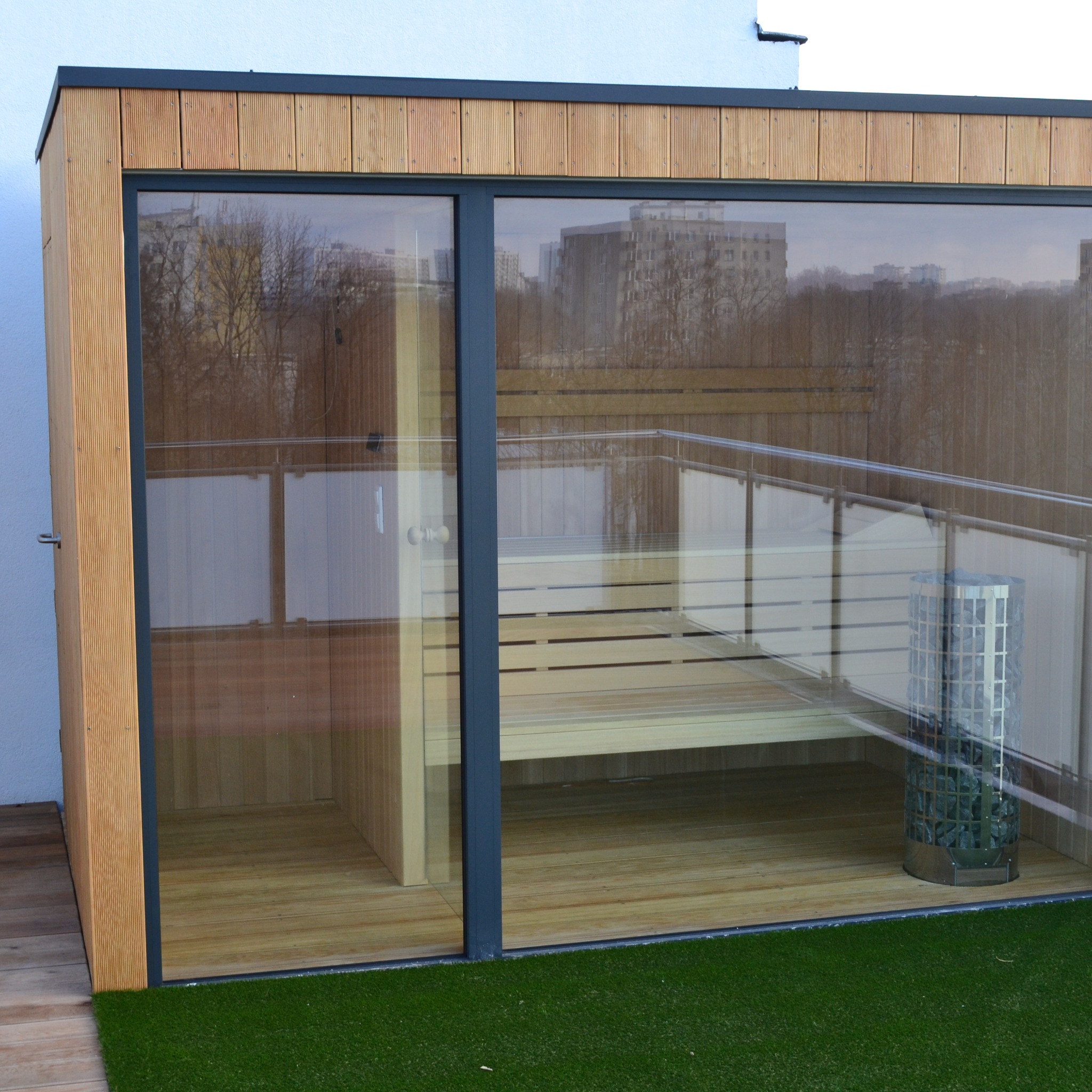Nowoczesna sauna zewnętrzna z przeszklonymi ścianami i drewnianym wykończeniem, widoczna ława i piec. Tło stanowi miejski krajobraz.
