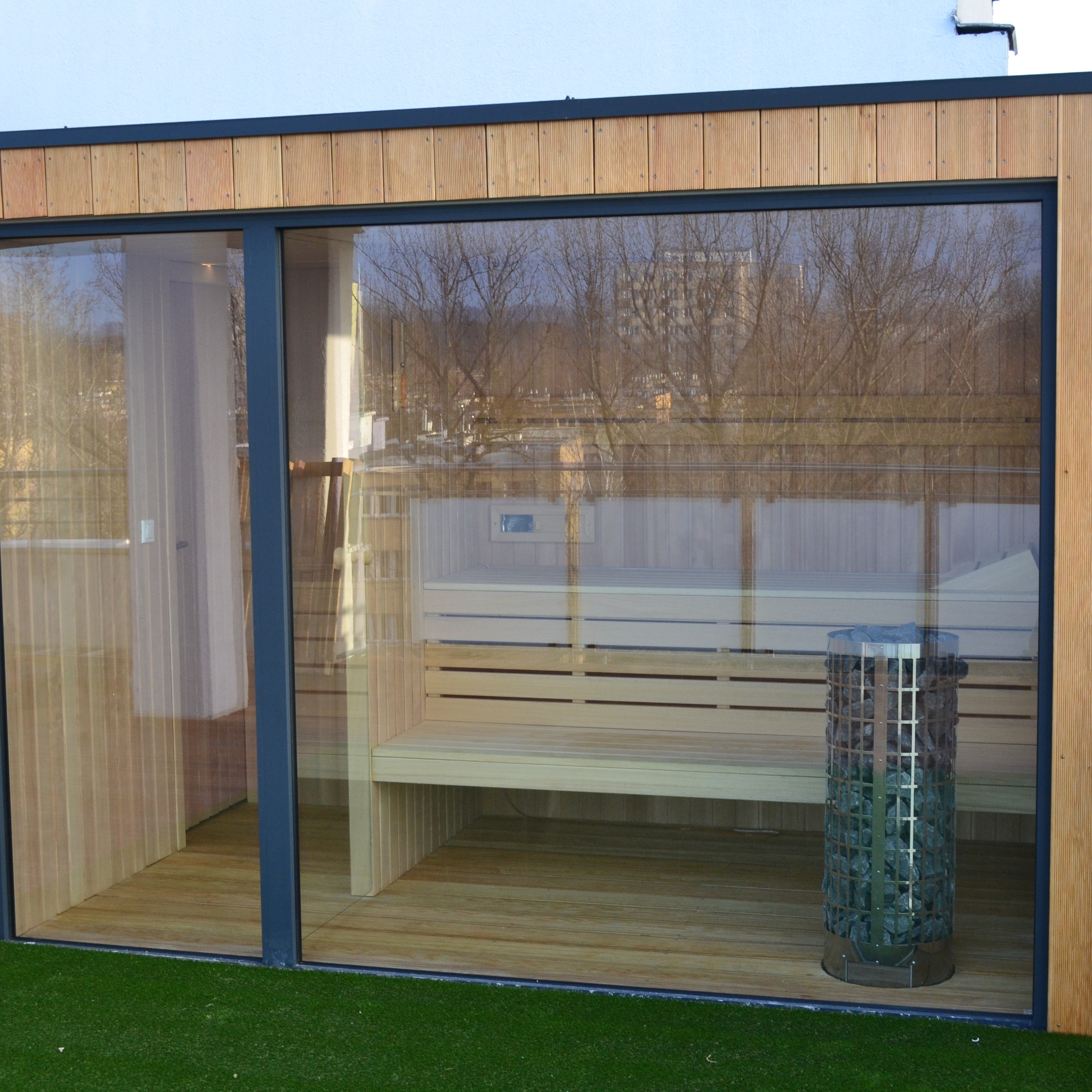 Nowoczesna sauna zewnętrzna z przeszklonymi ścianami, widoczne drewniane ławy i piec z kamieniami. Minimalistyczny design, jasne drewno, zielona trawa na zewnątrz.