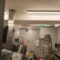 Wnętrze przemysłowej kuchni z nierdzewnymi urządzeniami, oświetleniem LED i oznakowaniem ewakuacyjnym. Widoczne elementy instalacji elektrycznej i wyposażenia.