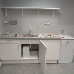 Biała szafka pod zlew z otwartymi drzwiczkami, widoczny bojler. Na blacie narzędzia, gniazdka elektryczne bez osprzętu. Na ścianie półki.