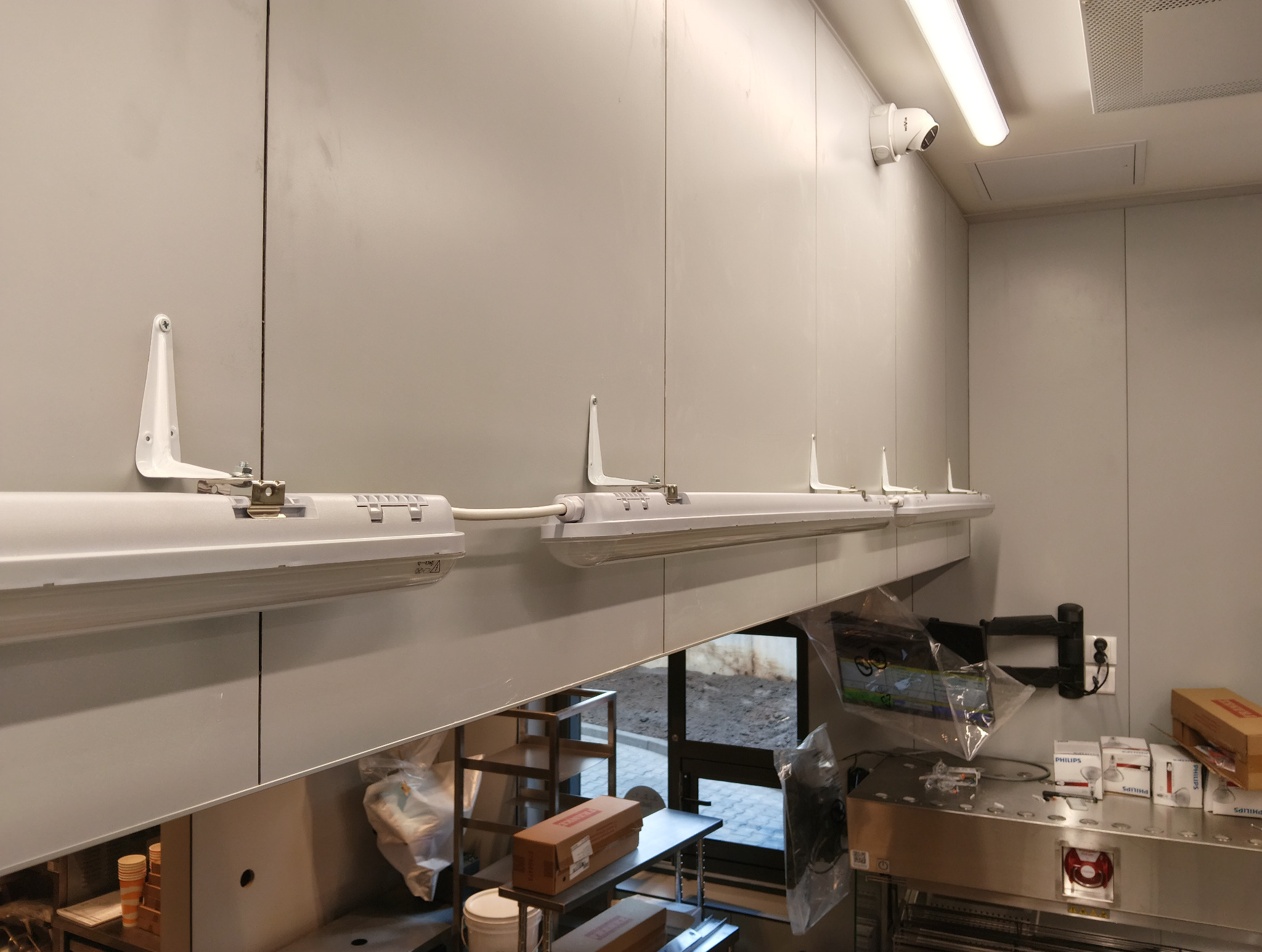 Zamontowane, podłużne lampy LED z białymi uchwytami na szarej ścianie. Widoczny fragment sufitu z kamerą. Metalowy blat i wyposażenie kuchni w tle.