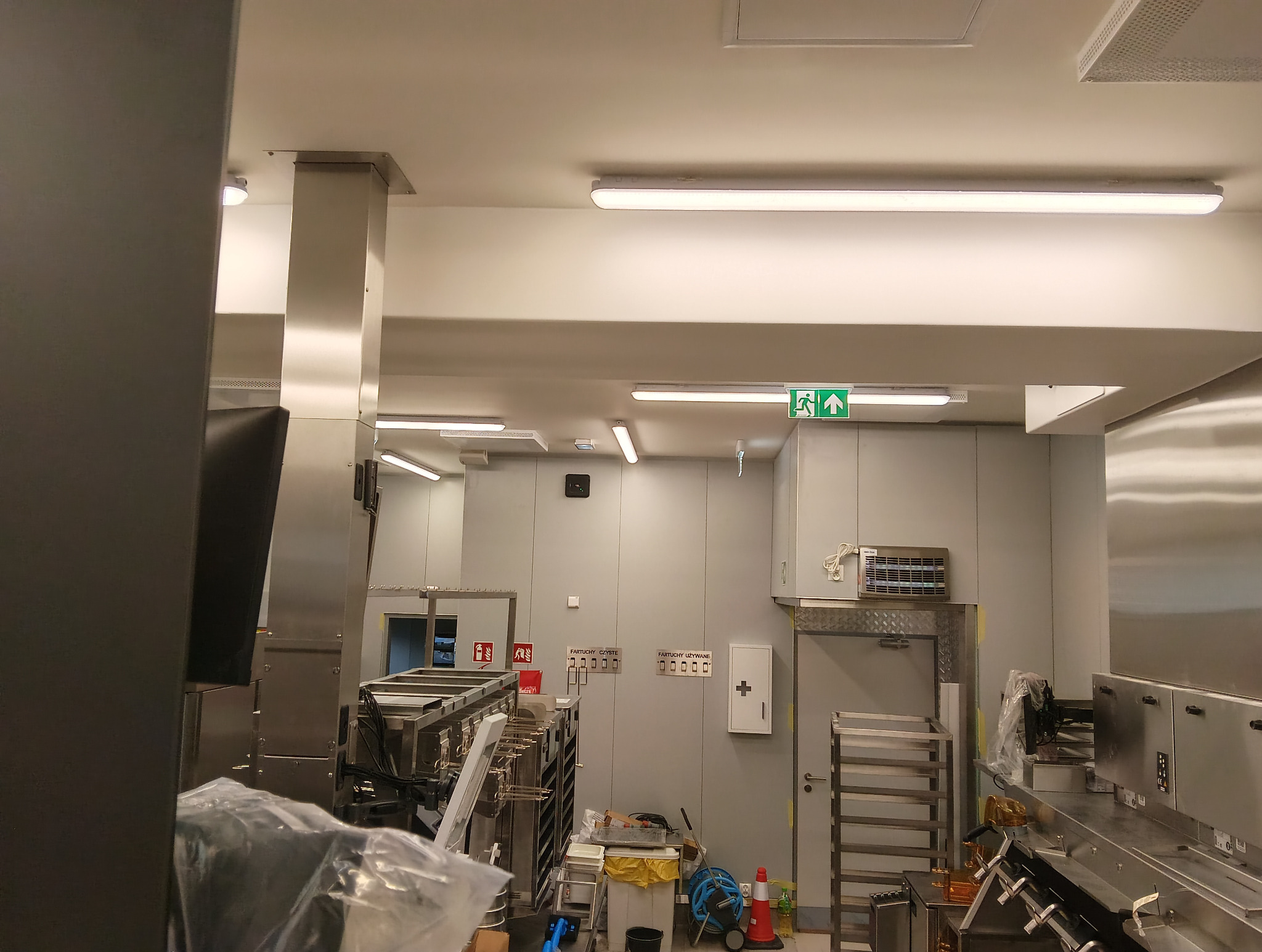 Wnętrze przemysłowej kuchni z nierdzewnymi urządzeniami, oświetleniem LED i oznakowaniem ewakuacyjnym. Widoczne elementy instalacji elektrycznej i wyposażenia.