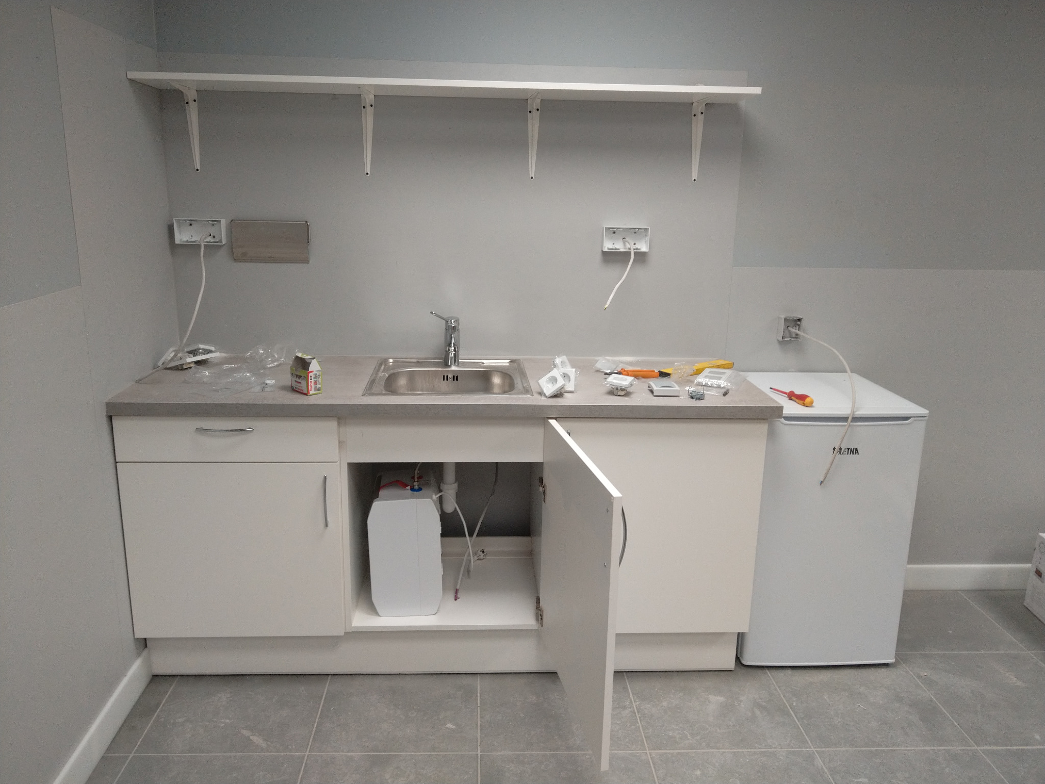 Biała szafka pod zlew z otwartymi drzwiczkami, widoczny bojler. Na blacie narzędzia, gniazdka elektryczne bez osprzętu. Na ścianie półki.