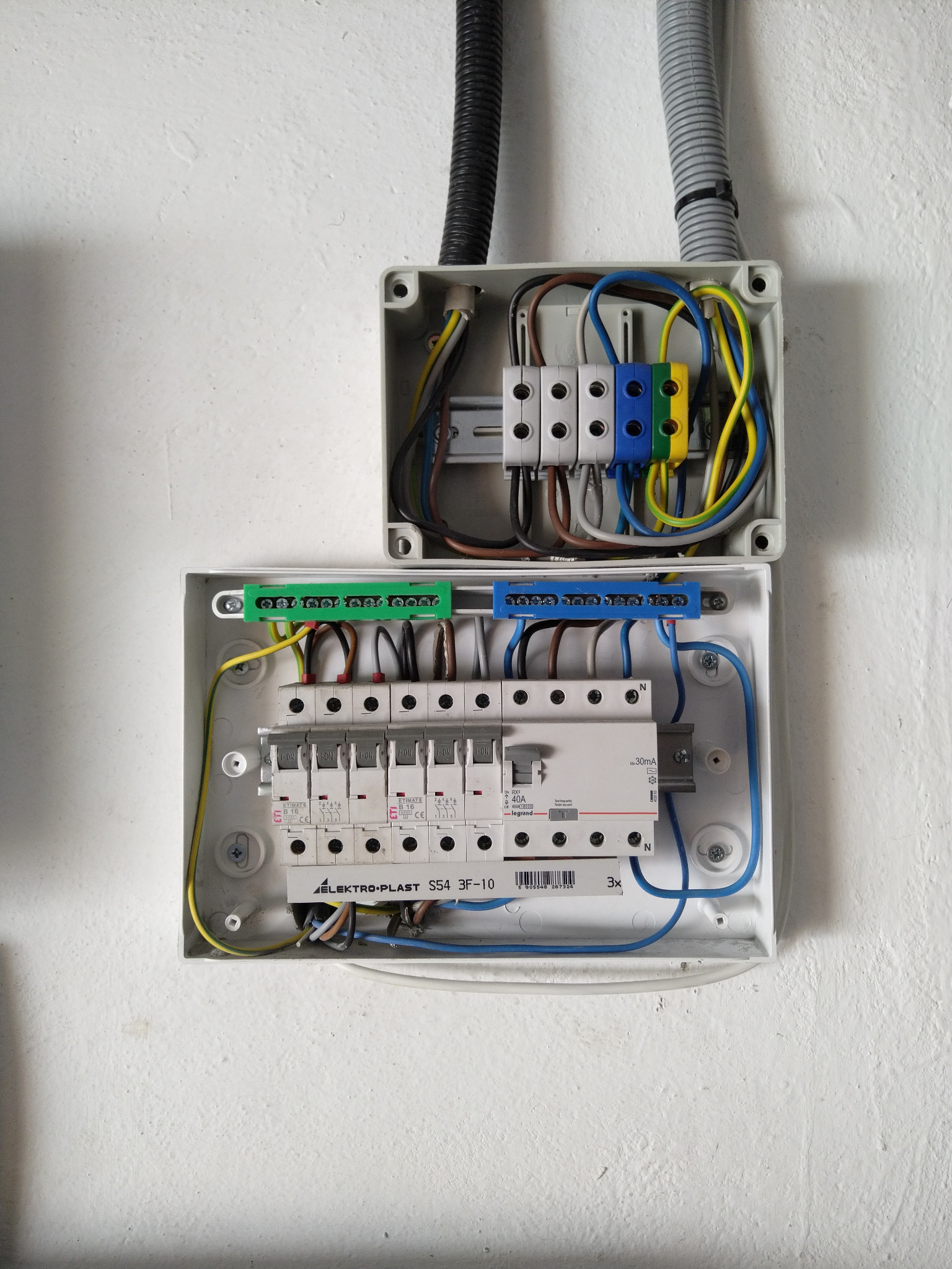 Otwarta skrzynka elektryczna z bezpiecznikami, widoczne kolorowe przewody i listwy zaciskowe. Instalacja na białej ścianie, przewody w peszlu.