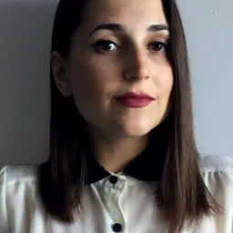 Portret kobiety o ciemnych włosach i brązowych oczach, ubrana w białą bluzkę z czarnym kołnierzykiem, na jasnoszarym tle. Usta pomalowane na ciemny kolor.